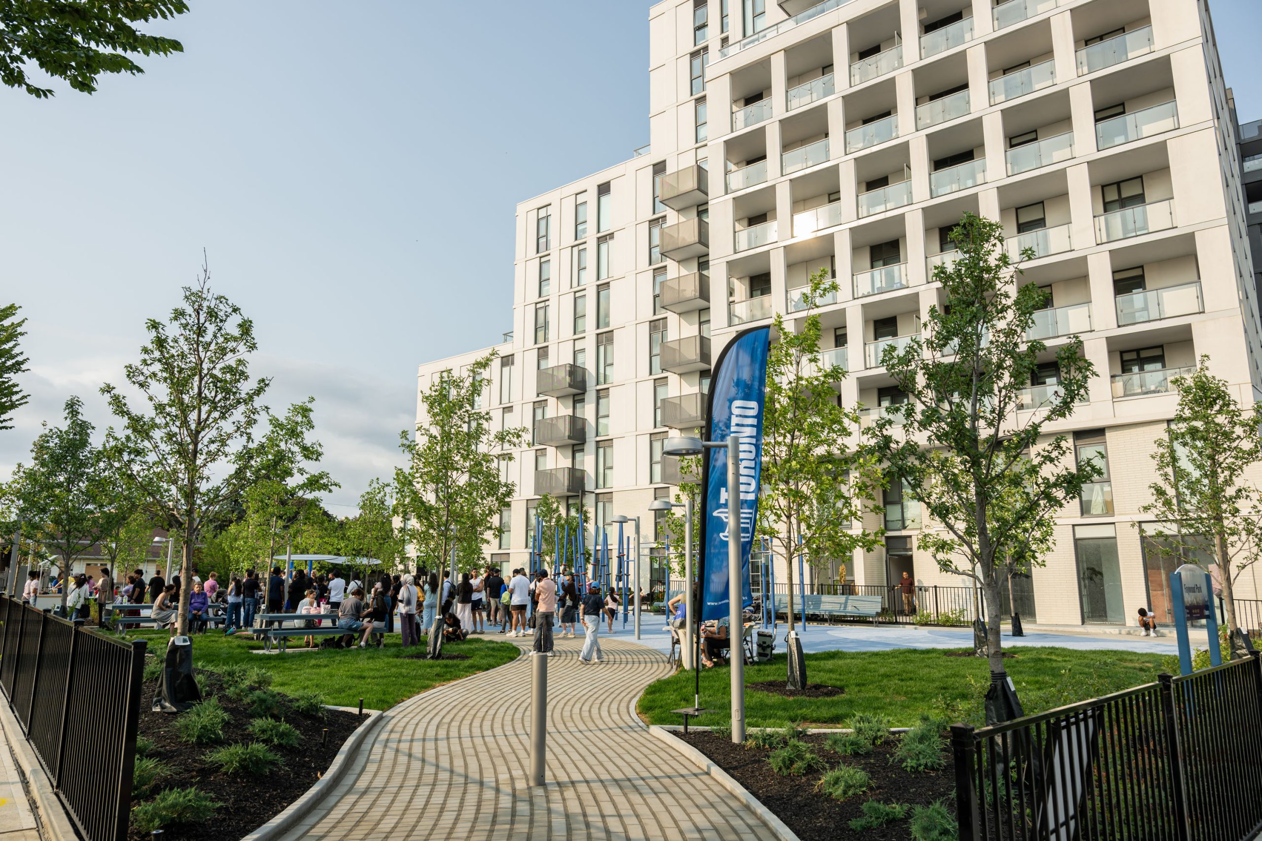  A paved pathway curves through a park, with grass on either side. The park includes picnic tables, benches and blue-coloured play structures. Community members are visible in the background enjoying the park.