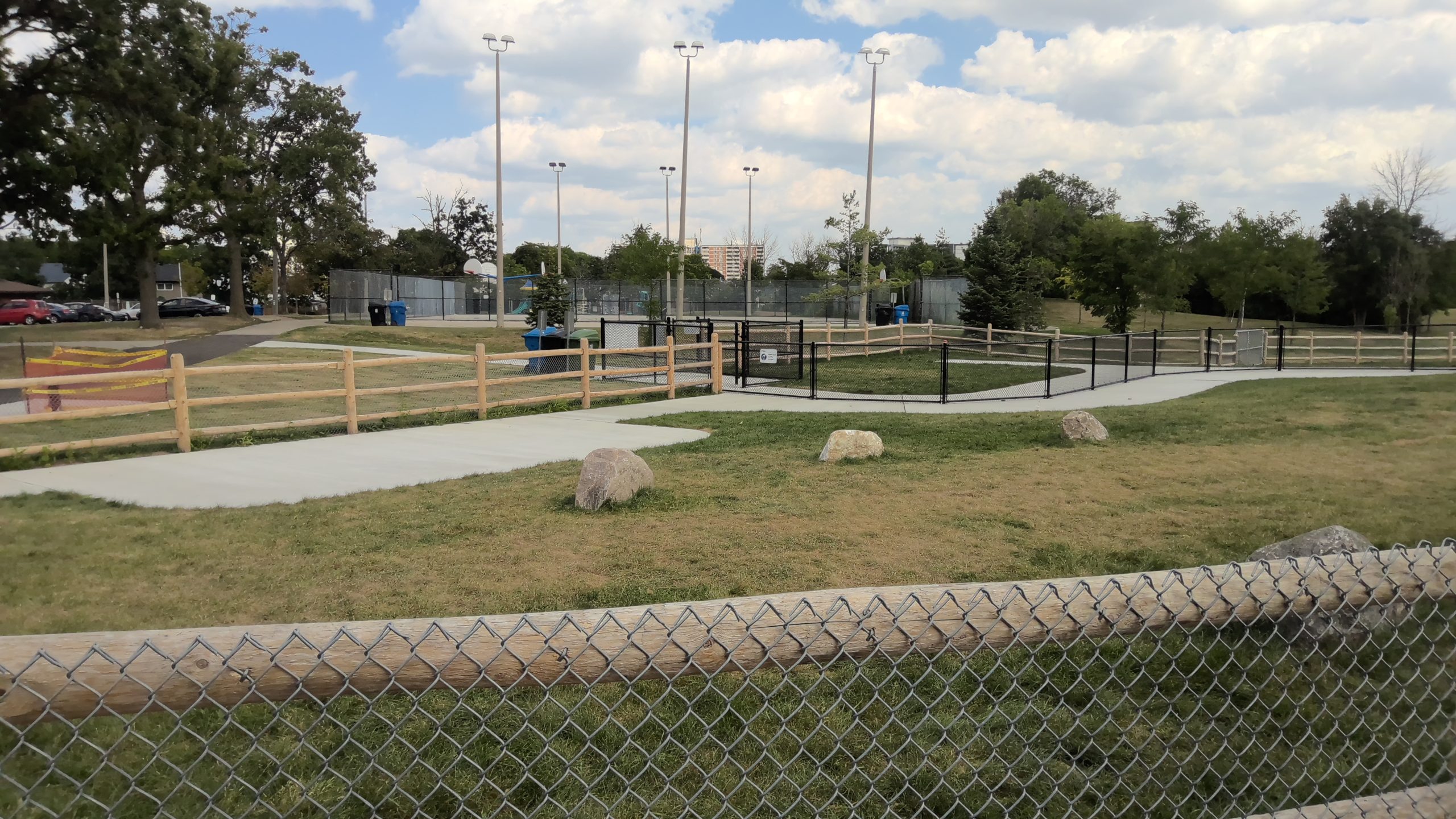 The new off-leash area featuring grass surfacing, small decorative boulders, paved pathways and both wooden post and paddle and black chain link fencing around the perimeter. The surrounding park has mature trees, grass lawns, pathways and basketball courts. Cars, houses and mid-rise buildings are visible in the distance.