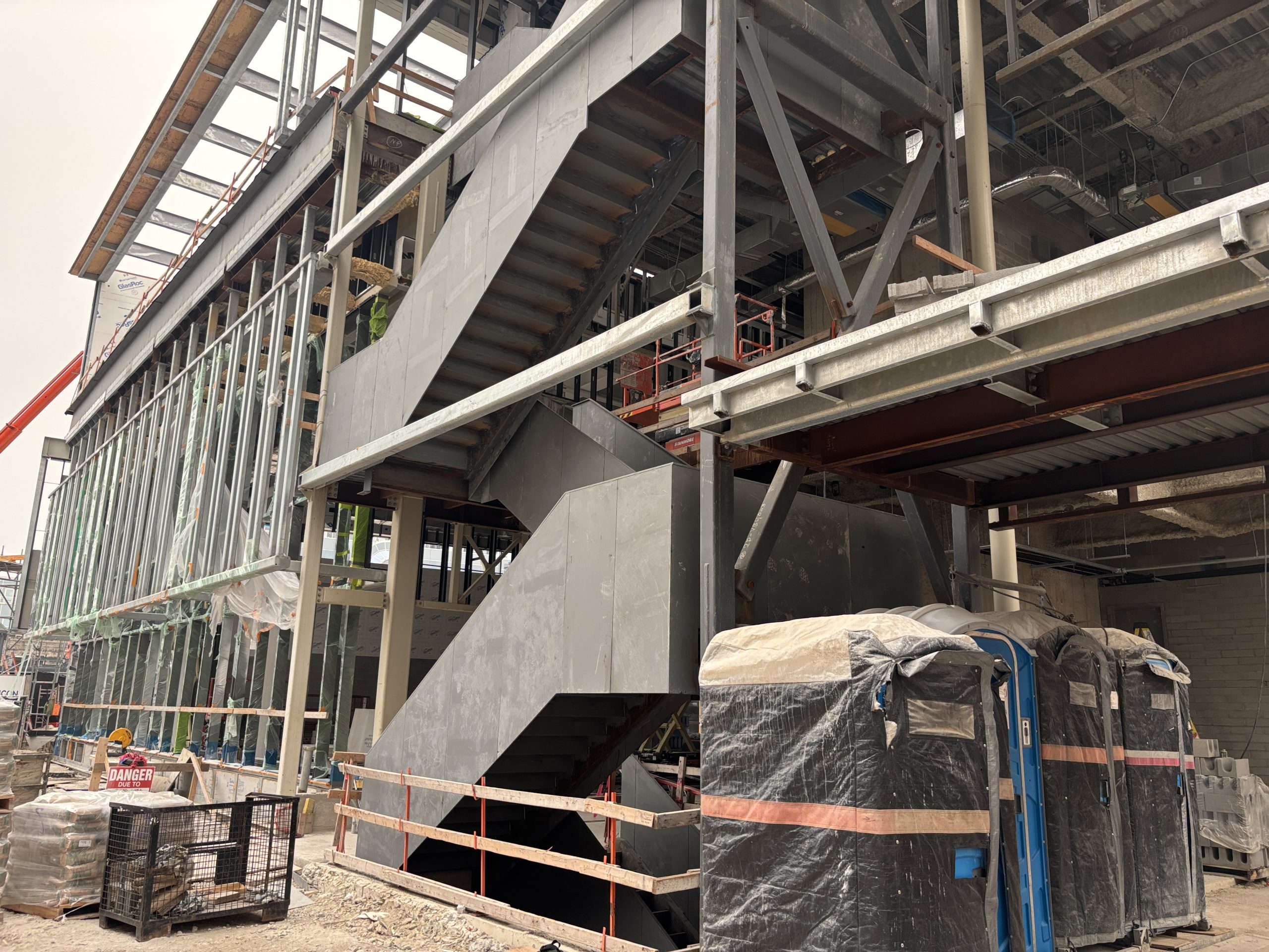 Exterior view of Davisville Community and Aquatic Centre from the side, showing a large multi-level staircase in progress. The building structure is under construction and comprised of exposed steel beams, columns and framing. Temporary safety railings, construction materials and portable toilets are visible on site.