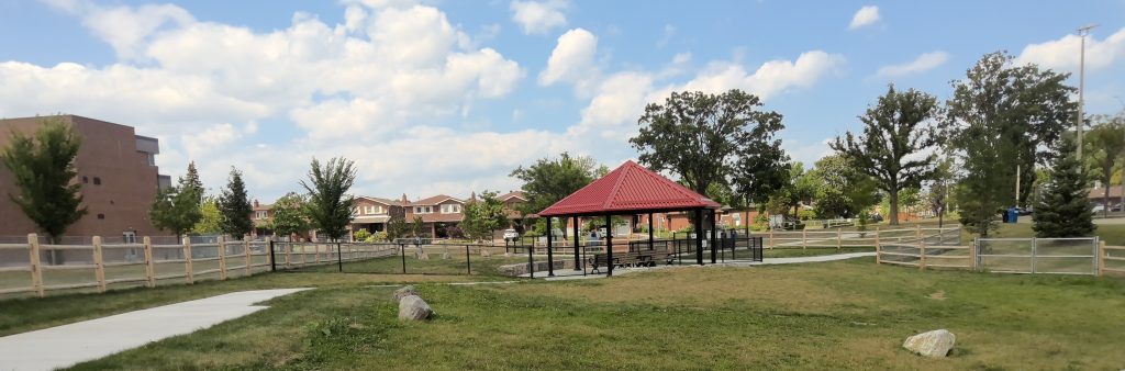An open grassy area surrounded by wooden post and paddle fencing. A paved pathway leads to a central shade structure with a red roof and benches underneath. A portion of the area is enclosed by black chain fencing. The area is bordered by a school building on the left, with residential houses visible in the distance.