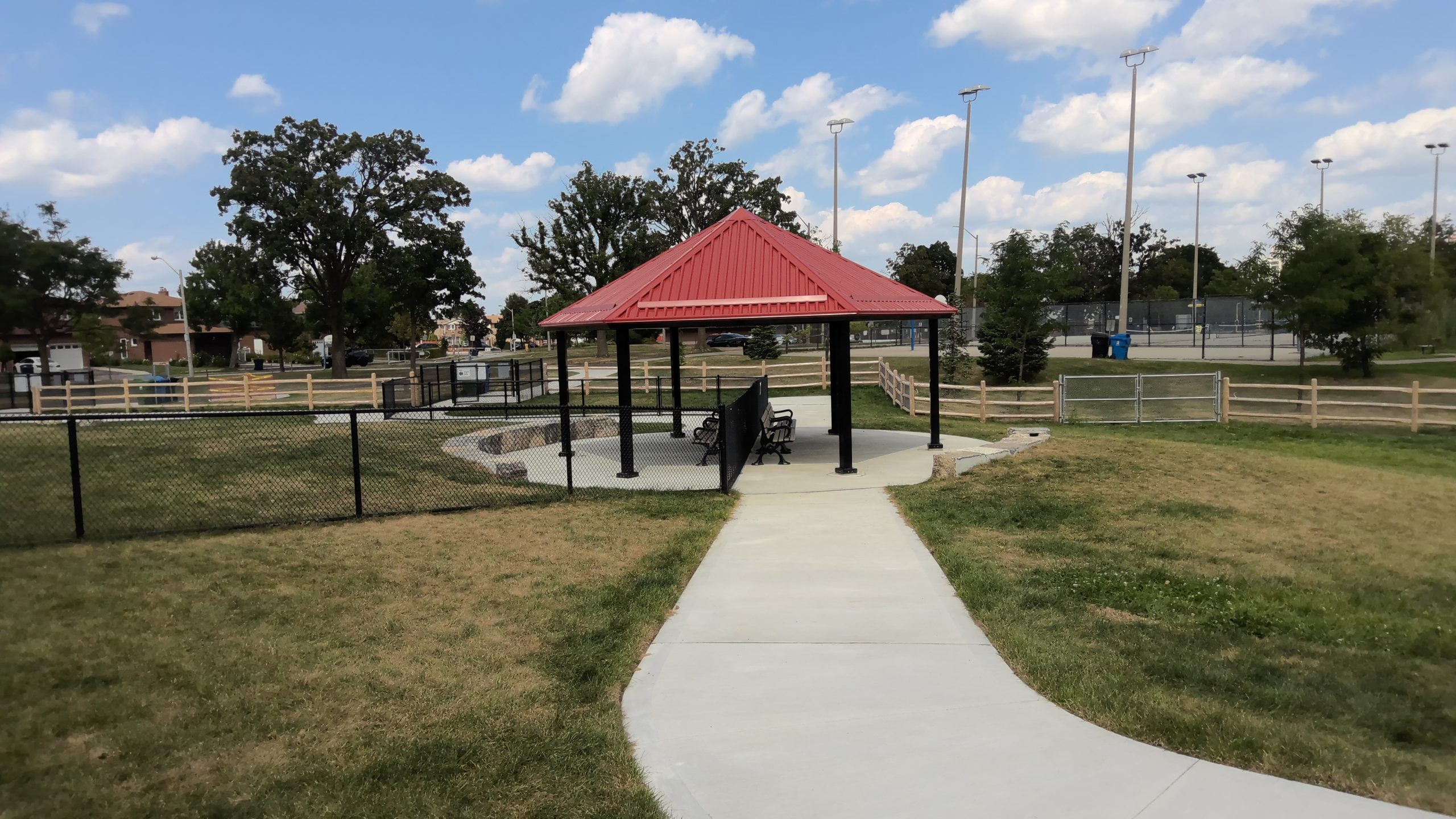 The shade structure in the new off-leash area featuring a pointed red roof supported by black columns on paved concrete. Benches are positioned underneath the canopy, while two armourstone seat walls curve around the shade structure. The surrounding off-leash area is fenced and surfaced with grass.