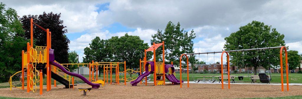 New Richmond Park playground looking north from the open lawn. New playground in is the middle ground with the senior and climbing structure to the left, junior structure in the center, and swings to the right. Soccer fields and baseball diamond are in the background.