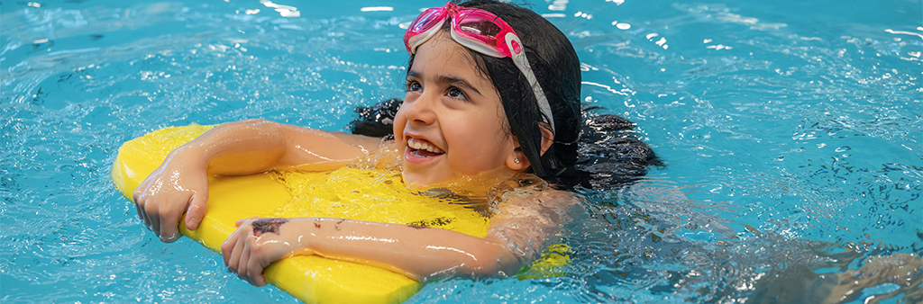 A girl swimming in a pool with a yellow kickboard and pink goggles.