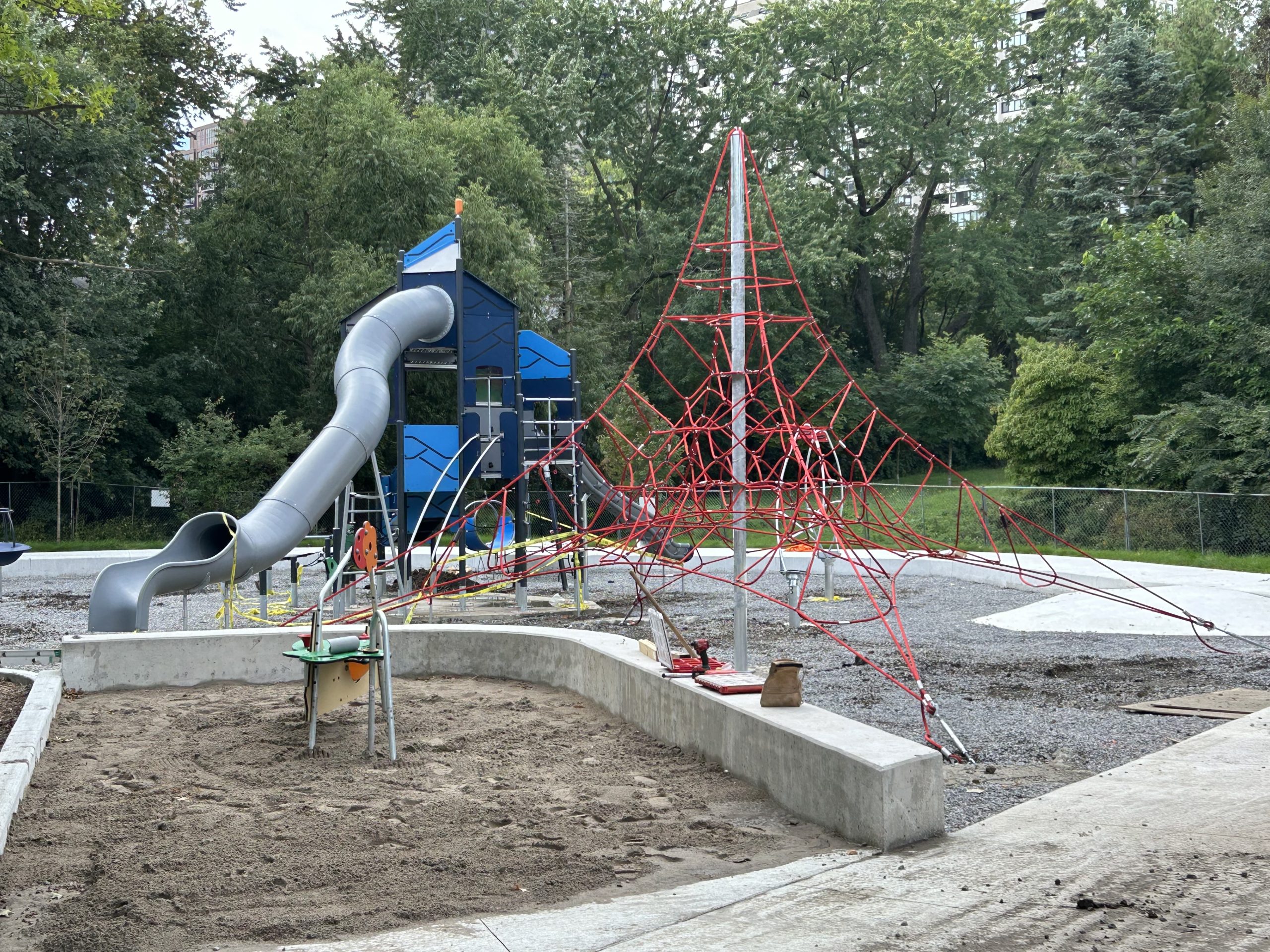 The newly installed senior play structure and net climber in Willowdale Park. The senior play structure includes a tall climbing tower and slides in a blue and grey colour scheme. The net climber is made of red cords interwoven in a large pyramid shape. Both structures are embedded in gravel and adjacent to a small sand play area. Construction tape and tools are visible on site.