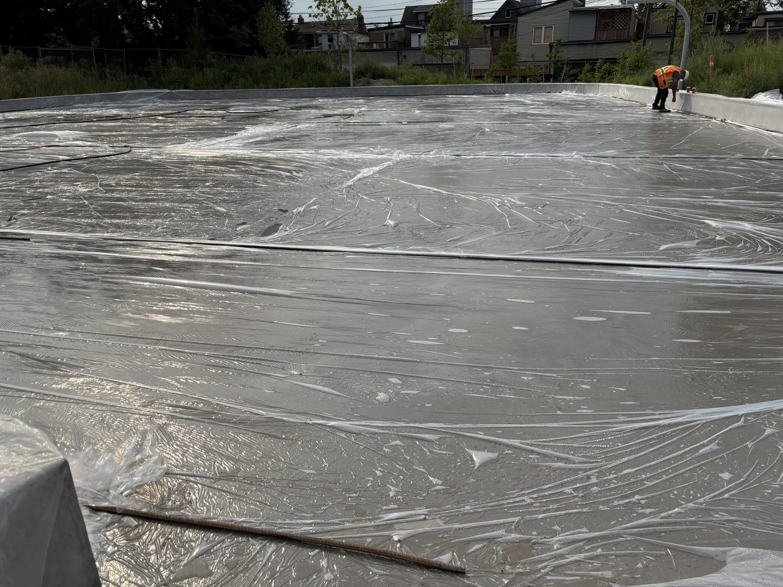 An outdoor rink under construction, showing a large concrete surface covered with protective plastic sheets. A construction worker is seen inspecting the raised concrete curb around the rink. Vegetation, fencing and residential buildings are visible in the background. 