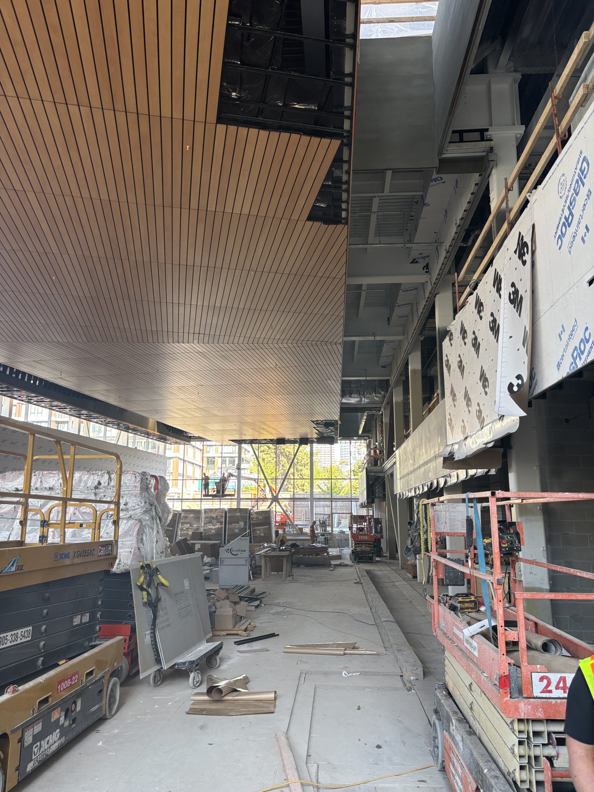 The interior of a large building under construction, featuring floor-to-ceiling windows along one side and an unfinished wall with exposed beams on the other side. Long wooden slats cover the ceiling, which is still in progress. The concrete floor is cluttered with stacks of boxes, construction materials and equipment, such as scissor lifts. Workers are visible around the open space.