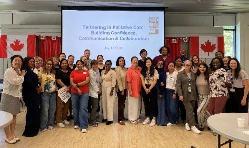 30 staff stand in front of a projector screen that says Partnering in Palliative Care: Building Confidence, Communication & Collaboration. Canadian Flags are in the background.