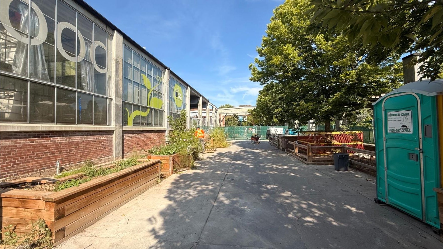 Construction activity along a wide concrete pathway by Barn 5 in Wychwood Barns Park. Planting beds line the edge where the pathway meets the building’s brick exterior, which has large glass windows. Mature trees line the other edge. Construction materials, a portable washroom and fencing are visible on the site, along with a construction worker.