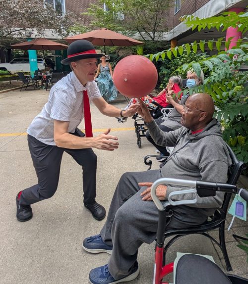 A busker wearing a rimmed bowler hat plays with a ball in front of a resident sitting on a wooden bench outdoors