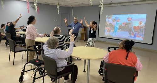 Wesburn Manor residents embark on a globetrotting adventure as part of the Travelogue program. Residents sit at tables while watching a projector screen and raising their hands as part of the activity.