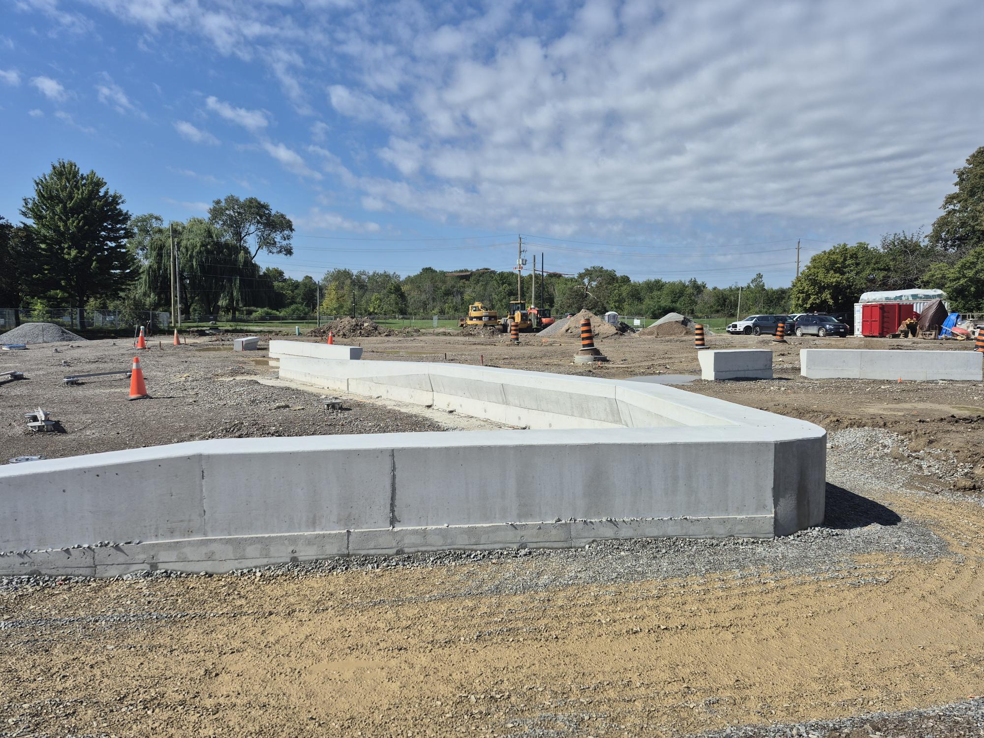 Construction site showing new concrete seatwalls in place. Orange cones, construction equipment and piles of soil are visible throughout the gravel site. Trees, open grass fields and power lines are visible in the distance.