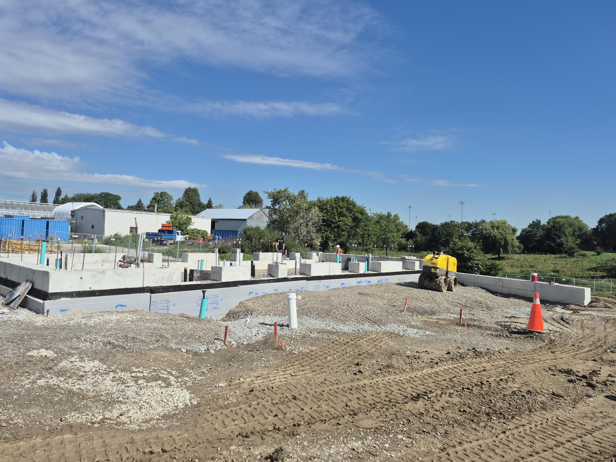 Construction site showing a rectangular concrete foundation in progress. The foundation extends slightly above the surrounding dirt and gravel ground. A small construction vehicle is parked next to the foundation. Grass fields, mature trees and greenhouse-style buildings are visible in the background.