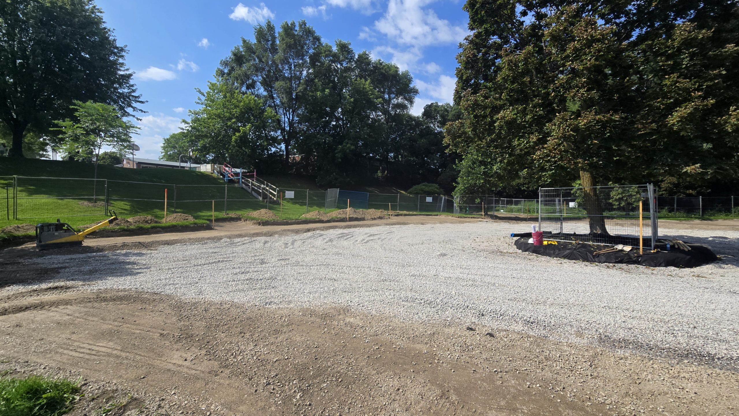Fenced construction site at Park Lawn Park. A mature tree is protected by fencing in the centre of a gravel area. The dirt ground along the outer edges is levelled. Beyond the construction site, grassy expanses, trees and a set of stairs are visible. 