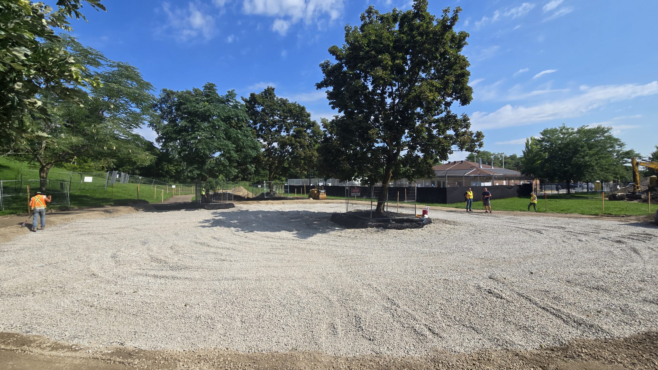 Construction site at Park Lawn Park, showing progress for the new playground. The large circular area is surfaced with gravel and includes a large tree at the centre. Workers are visible around the site, along with construction equipment. 