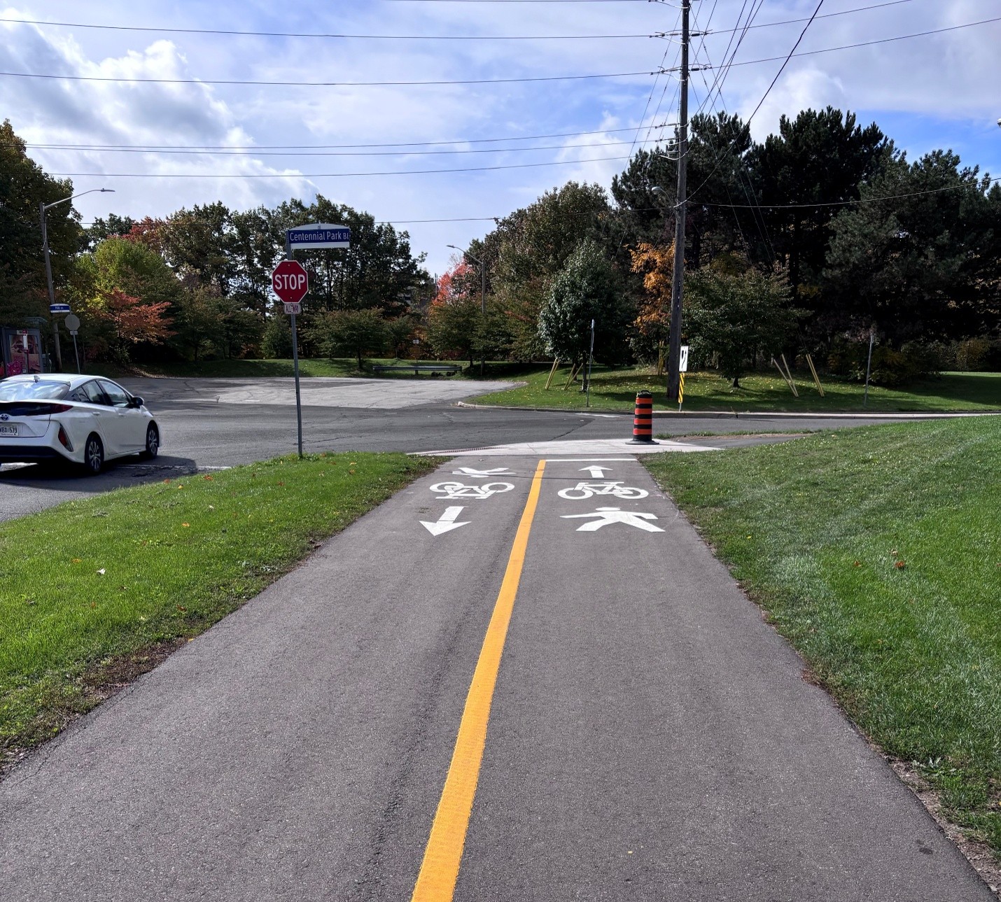 A paved multi-use trail with a yellow centre line and white pedestrian and cyclist direction markings. The path is bordered by grass and connects to a crossing at Centennial Park Boulevard.