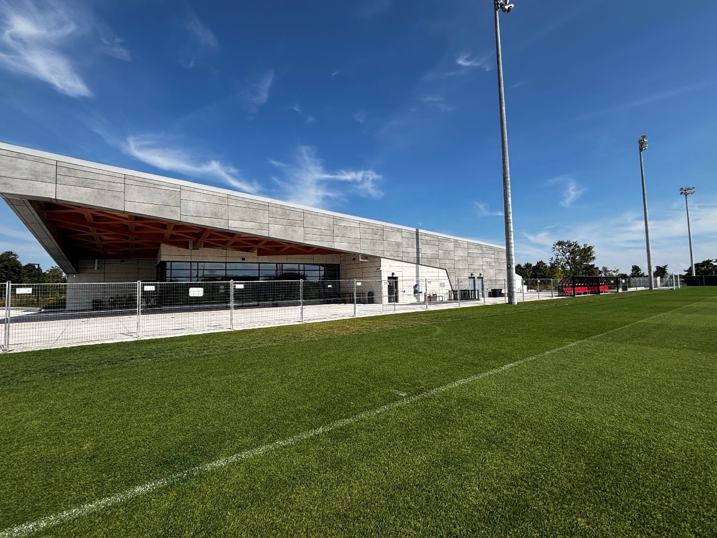 View of a building beside a soccer field. The building features a large covered canopy with exposed timber ceiling elements. Tall light poles and perimeter fencing are visible along the field.