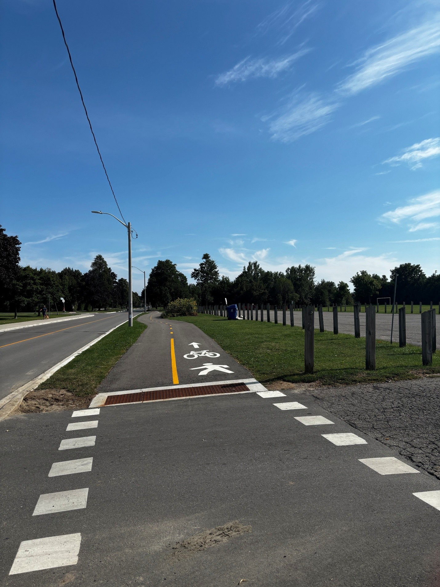 A paved multi-use trail marked for pedestrians and cyclists beside a roadway. Grass borders both sides of the path, with wooden posts lining an open field to the right and trees in the distance.