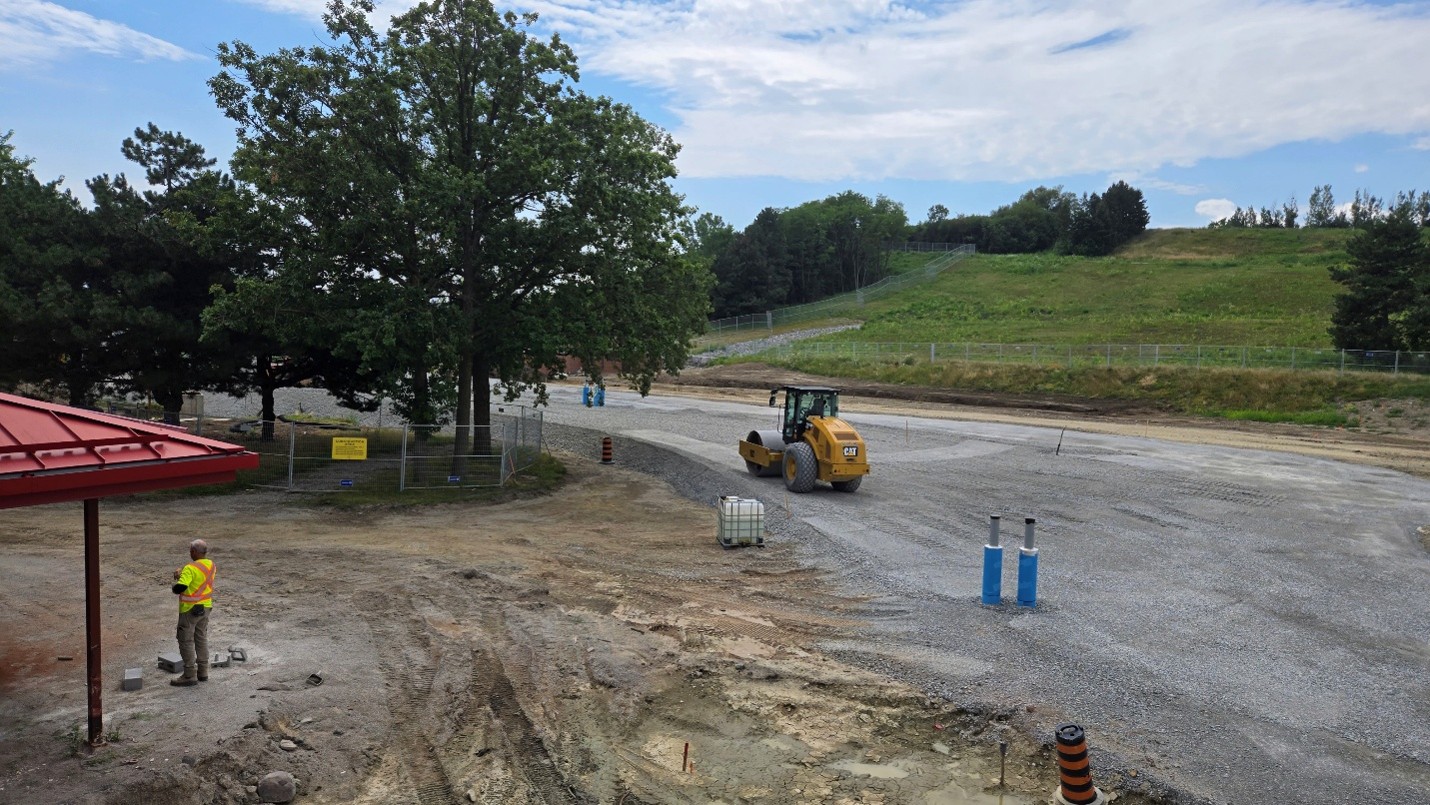 Construction site showing a machine operating on a large gravel surface. Blue utility pipes extend from the ground. A fenced grove of mature trees stands to the left, beside a red-roofed structure, while a construction worker stands nearby.