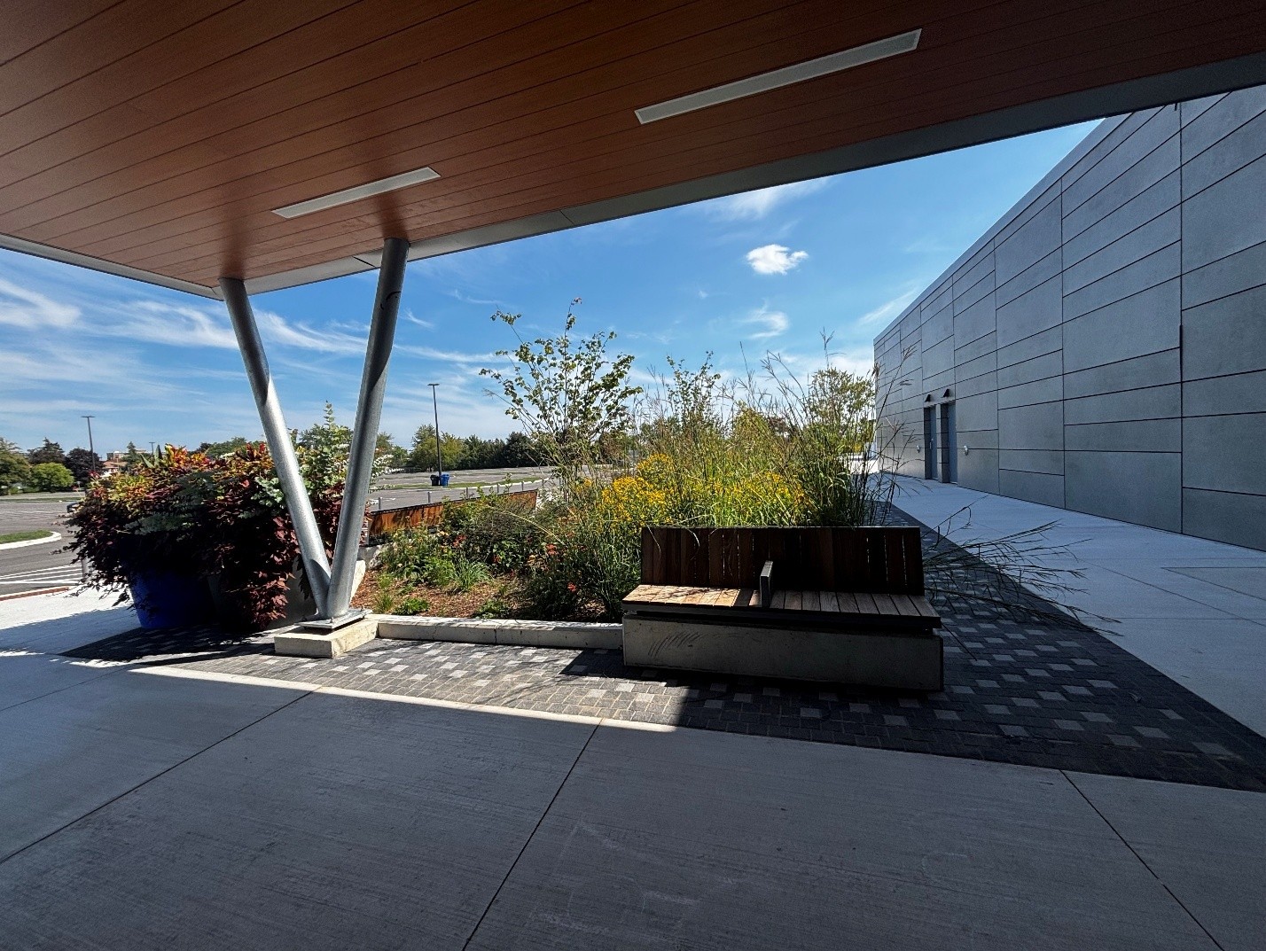 Outdoor paved area beside a building, featuring landscaped planting beds, wooden bench seating and wide concrete walkways. A covered canopy supported by angled columns provides shade, while a parking lot and trees are visible in the background.