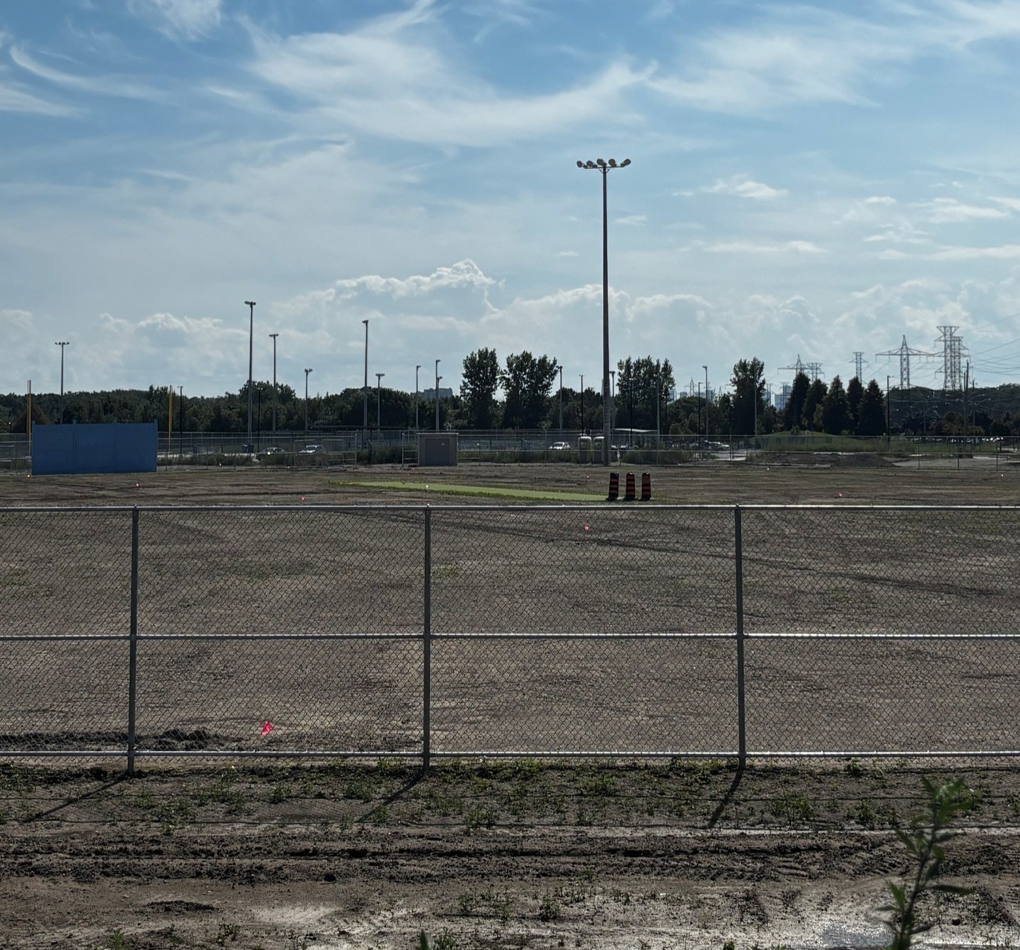 View of a fenced construction site with a large, graded gravel surface in the foreground. Several tall light poles stand across the site, with a blue wall structure visible to the left. Trees line the background, with hydro towers and power lines visible in the distance.