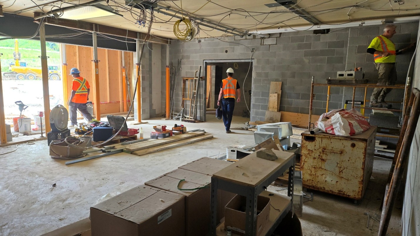 Interior view of a building under construction. Concrete block walls, exposed ceiling wiring and unfinished door openings are visible. Construction workers are at work in the space, which contains construction materials and equipment. 