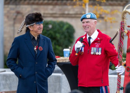 Two people stand outdoors, one wearing a feathered headdress and the other holding a microphone and a decorated staff while wearing a red Métis Nation of Ontario jacket.