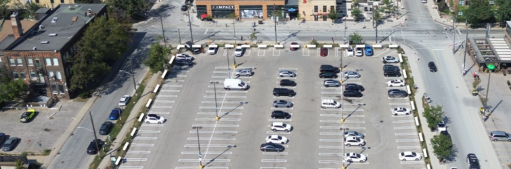 Aerial view of the new park site, showing the existing surface parking lot. Surrounding streets, (Hana Avenue to the East, Liberty Street to the North and Atlantic Avenue to the West), and multi-storey commercial and residential buildings visible on all sides.