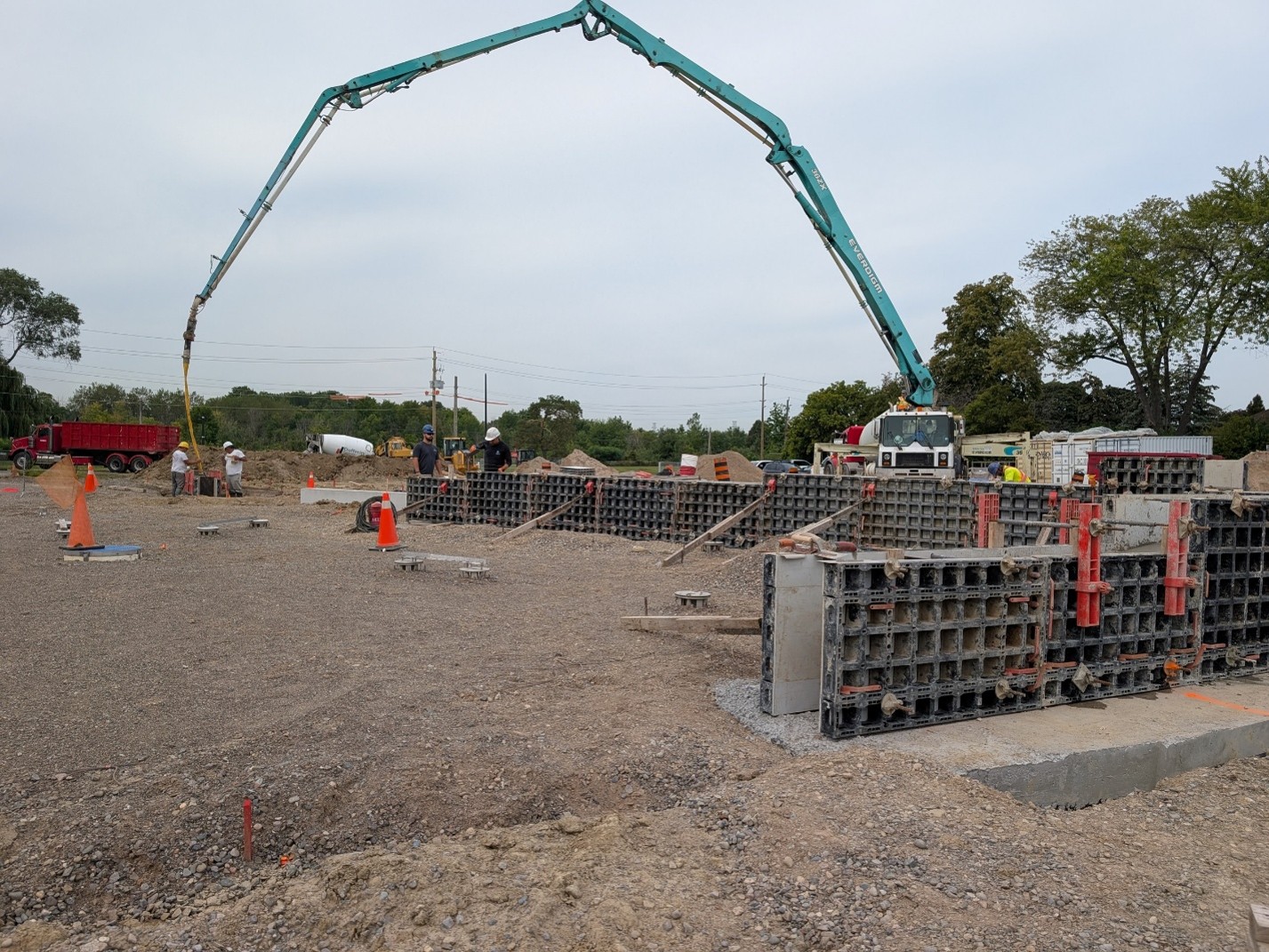 Construction site where concrete is being poured into rectangular forms. A truck with an extended arm arches across the site to where two workers are filling the concrete forms. Other construction equipment and machines are visible around the site, as trees and power lines span the background.