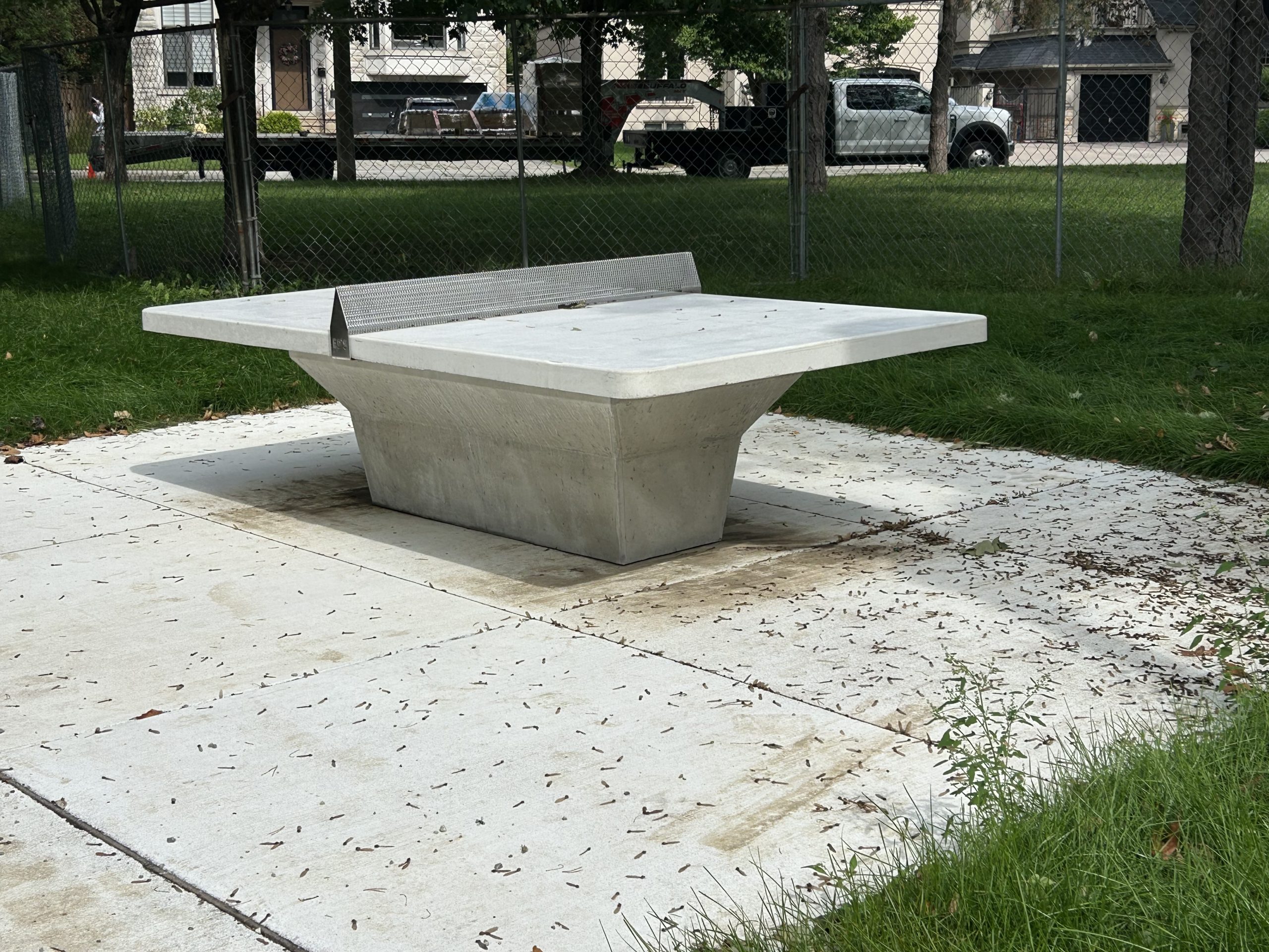 A newly installed concrete ping pong table with a metal net in Willowdale Park. The table is set on a concrete pad in a grassy area surrounded by fencing. In the distance, trees, houses and vehicles are visible.