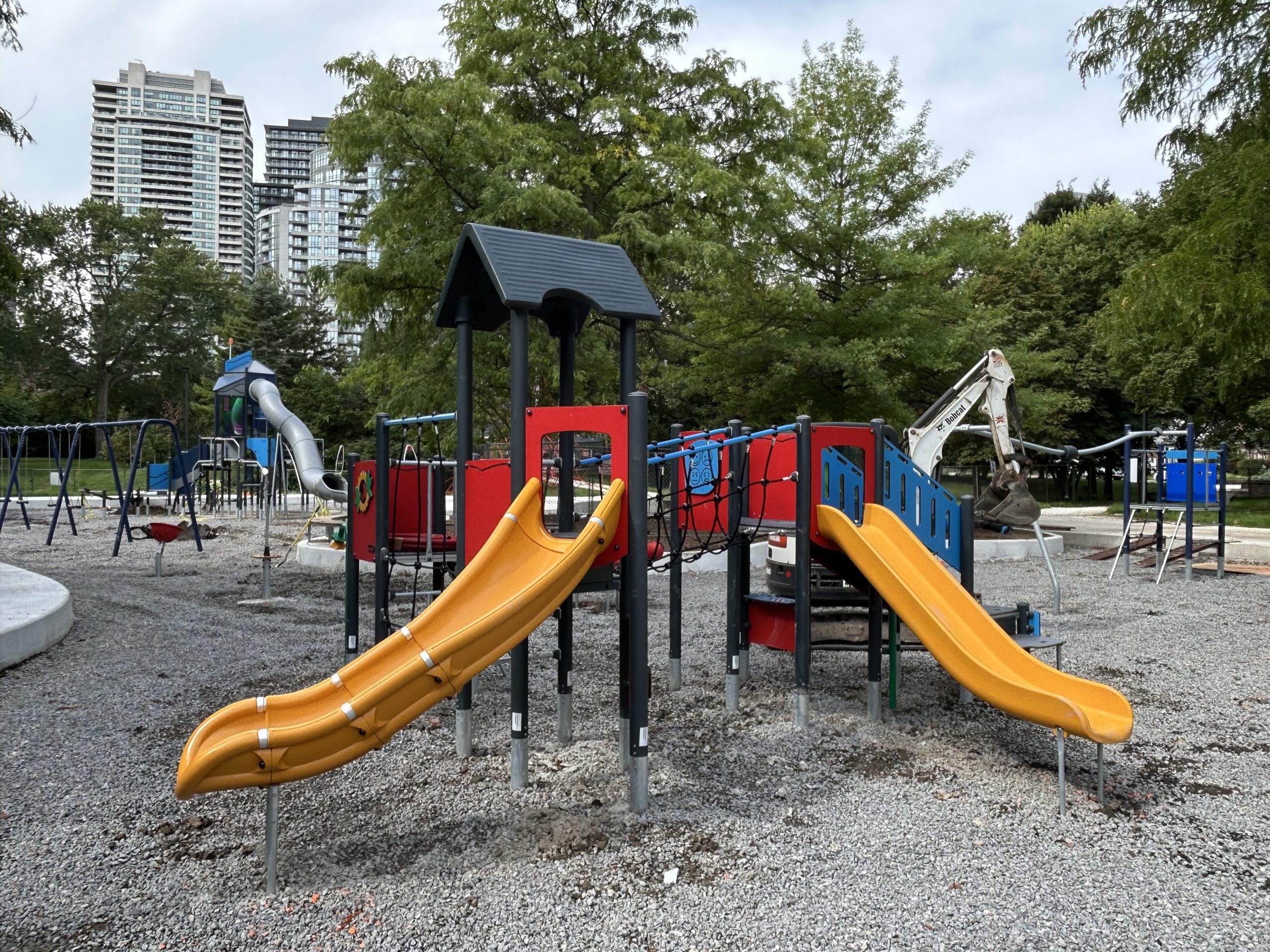 The newly installed junior play structure in Willowdale Park, with two slides and multiple climbing options in a red, yellow, blue and black colour scheme. The structure is embedded in gravel and surrounded by other equipment, such as a swing set, seated spinner and senior play structure. An excavator is visible on site. Trees and mid-rise buildings line the background. 