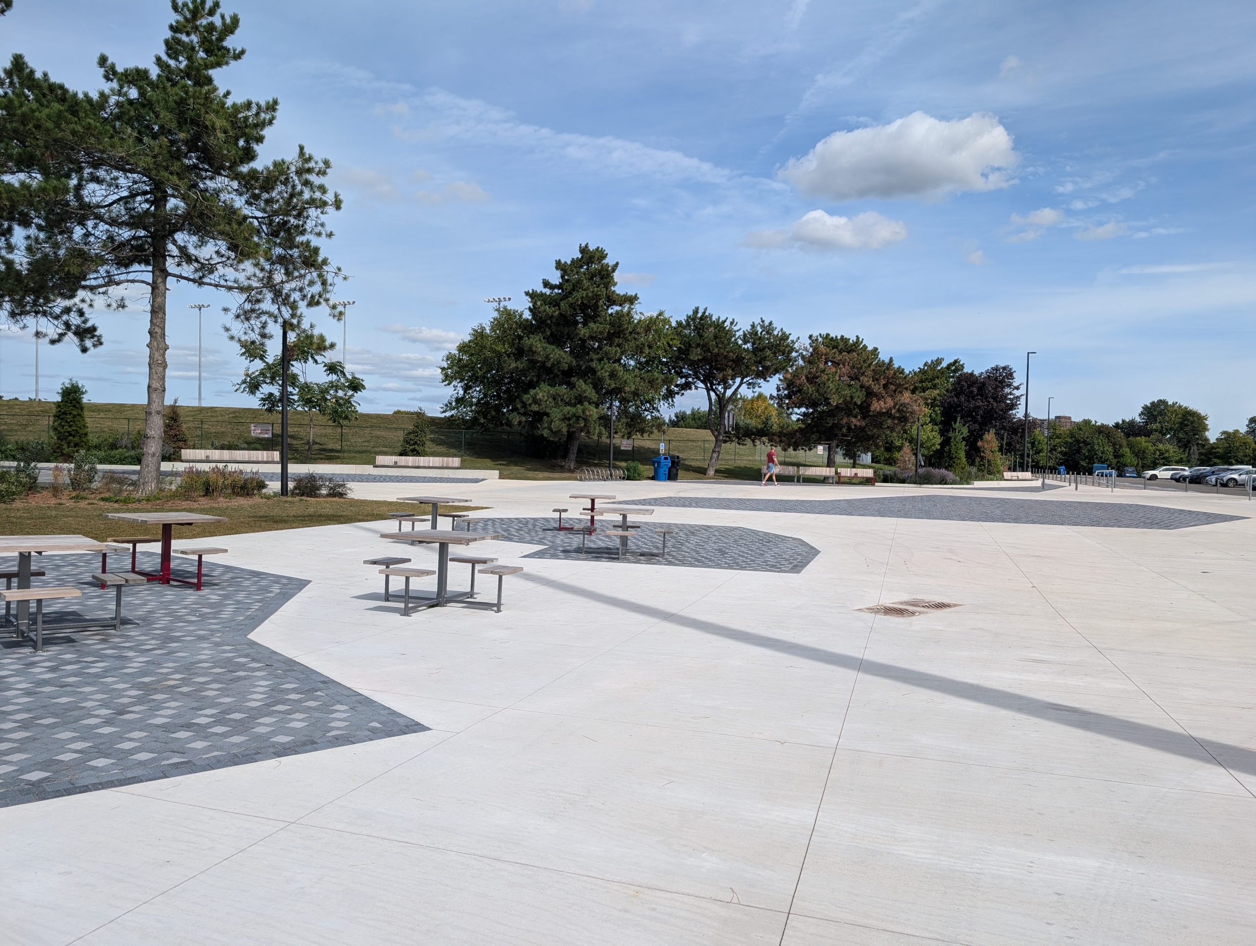 A large paved plaza featuring a landscaping bed and several square tables with stools attached at all four sides. Mature trees, tall light poles and grass fields line the background. 