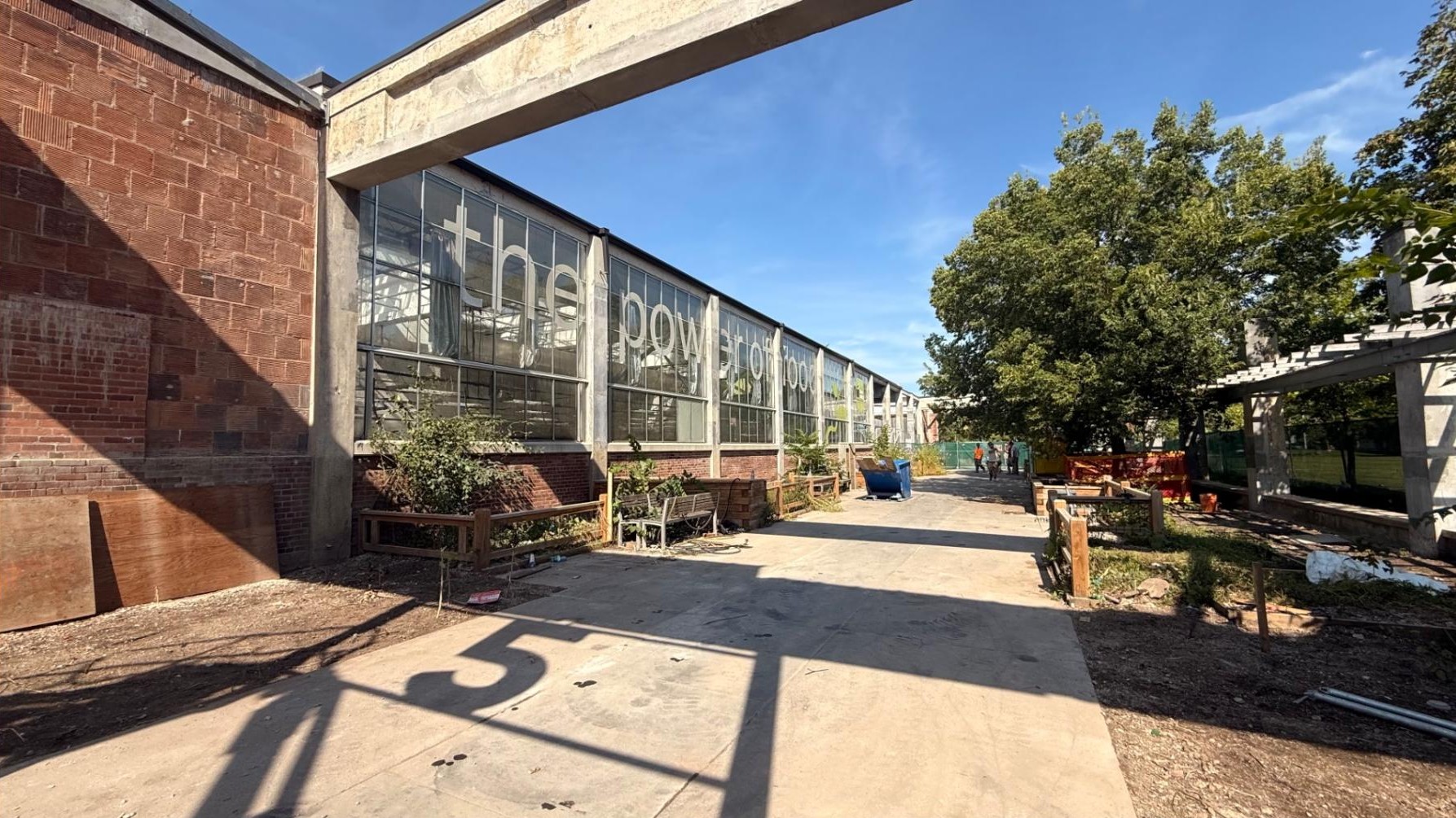 Exterior view of Barn 5 in Wychwood Barns Park, a large brick-and-glass building with “the power of food” displayed across its windows. A wide concrete pathway along the path is lined by planter boxes, benches and mature trees. Construction materials and fencing are visible around the area.