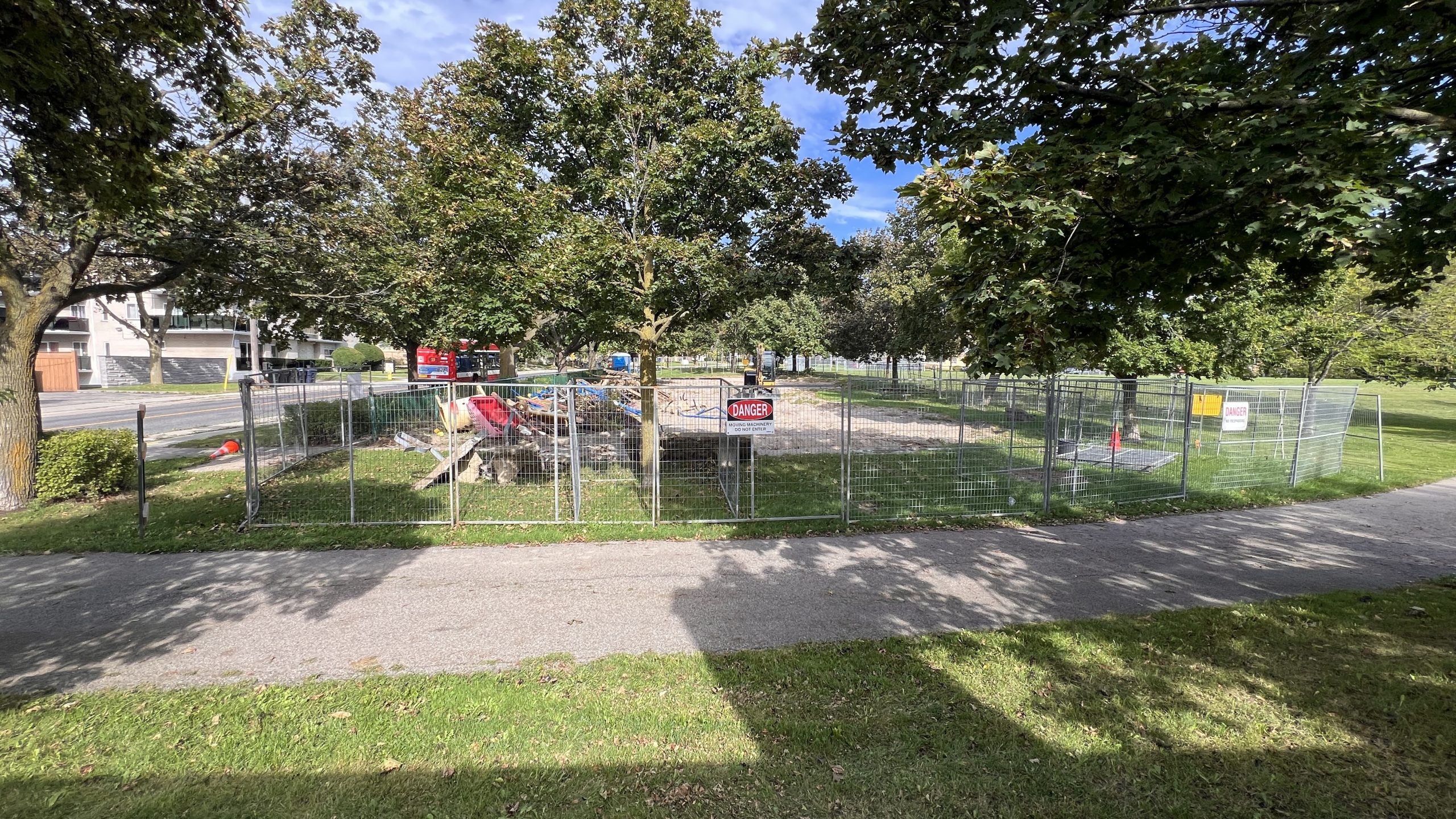 Fenced construction site in a park, showing demolition underway. Dismantled and scrap materials are piled on one side of a sand lot, where a construction machine is parked. The surrounding park features open grass fields and mature trees.