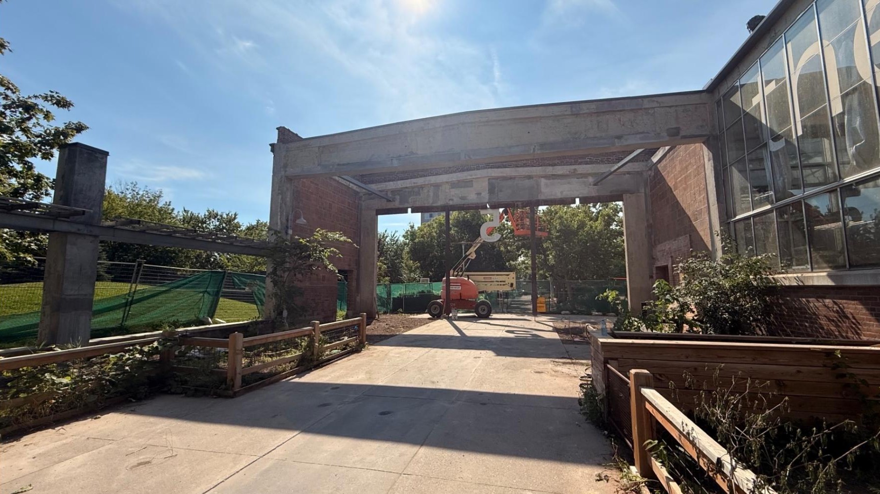 Construction activity at the entrance of Barn 5 in Wychwood Barns Park, a brick building with glass windows, an open canopy structure and a concrete pathway lined with planting beds. A lift is positioned beneath the canopy, with fencing visible around the building. The surrounding area includes grass lawns and trees.