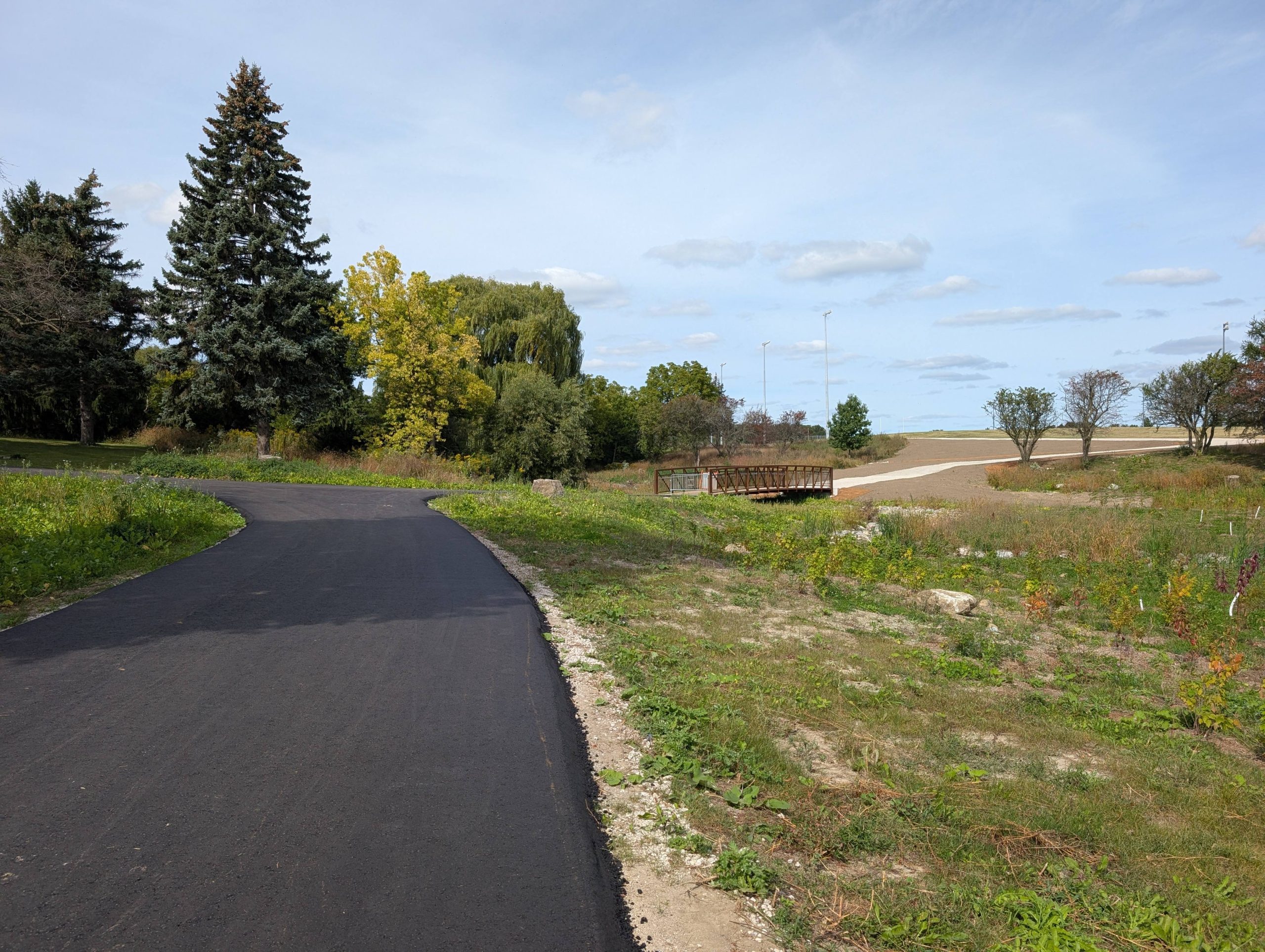  A newly paved black asphalt walking path curves through the naturalized Elmcrest Creek landscape towards a new wooden pedestrian bridge crossing the creek. Mature trees are visible in the surrounding area.