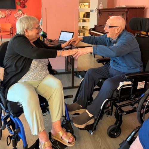 Two residents in wheelchairs touch hands while listening to music at Lakeshore Lodge.