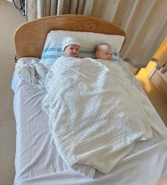 Two baby dolls are placed under a blanket in a bed with their little heads resting on a pillow.