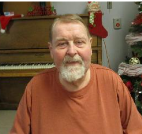 Stephen Oliver sits in front of a piano wearing a orange shirt and smiles