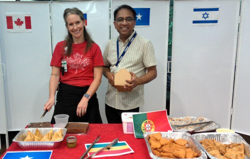 Two staff pose for a photo behind a table laden with food. A Canadian Flag is on the wall in the background and the table is decorated in a red table cloth.