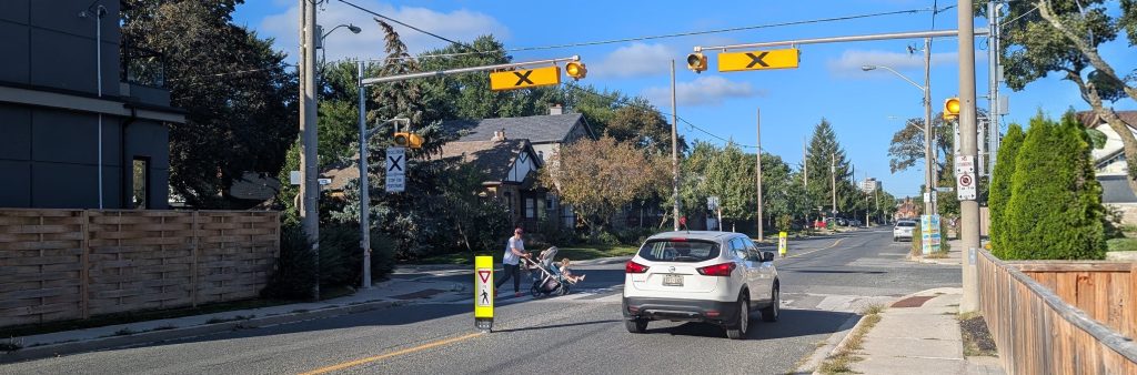 A pedestrian crossover crossing Mortimer Ave at Dewhurst Blvd. A person pushing a stroller is crossing the road with a driver yielding before the crossover. The pedestrian crossover has been enhanced with a flexible in-road sign in advance to increase visibility.