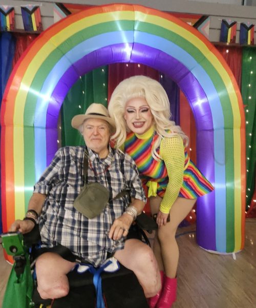 A resident of CWT sits in their wheelchair posed beside a professional drag queen entertainer beneath a rainbow archway.