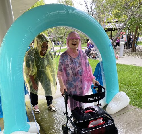 A resident at Bendale Acres stands with her walker under a sprinkler wearing a rain poncho.