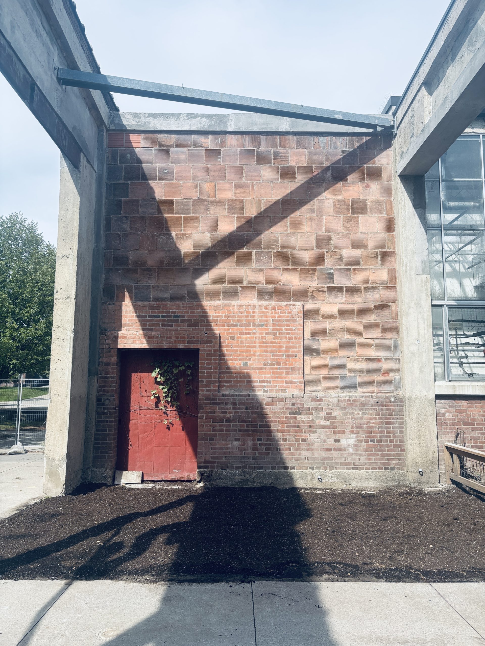 The brick façade of a large building, framed by concrete columns on all sides and a steel beam overhead. A red door in the corner is partially covered by vines. A rectangular bed of soil lies immediately at the door, surrounded by concrete paths.