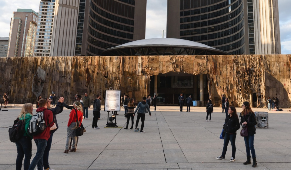 People walking across Nathan Phillips Square and City Hall covered in an artwork. Rotunda and towers behind.