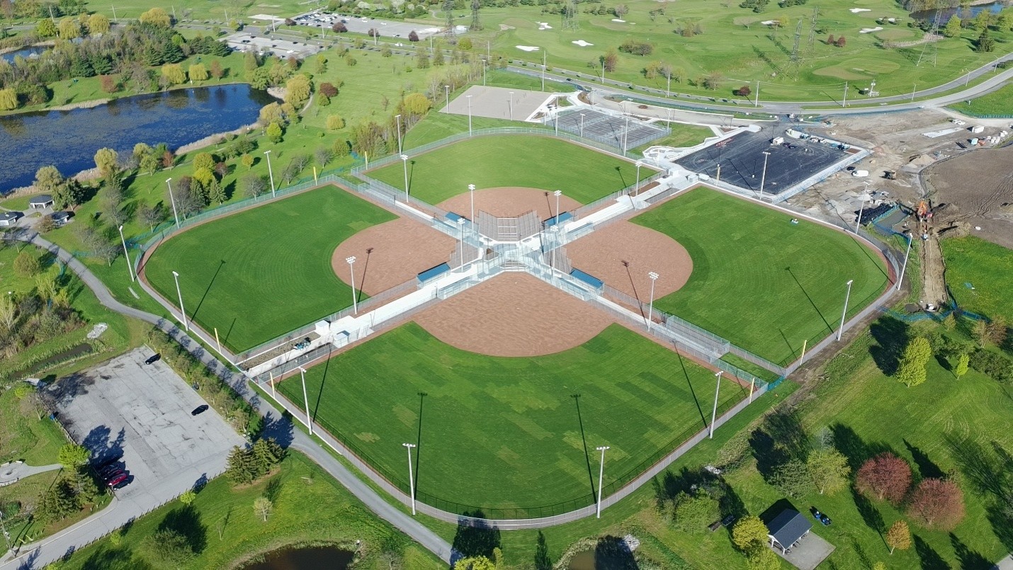 Aerial view of a four-diamond baseball complex with a central shared hub, fenced playing fields, dugouts, bleachers and tall field lights. The sports facility is surrounded by green parkland, walking trails, ponds, parking areas and roads.