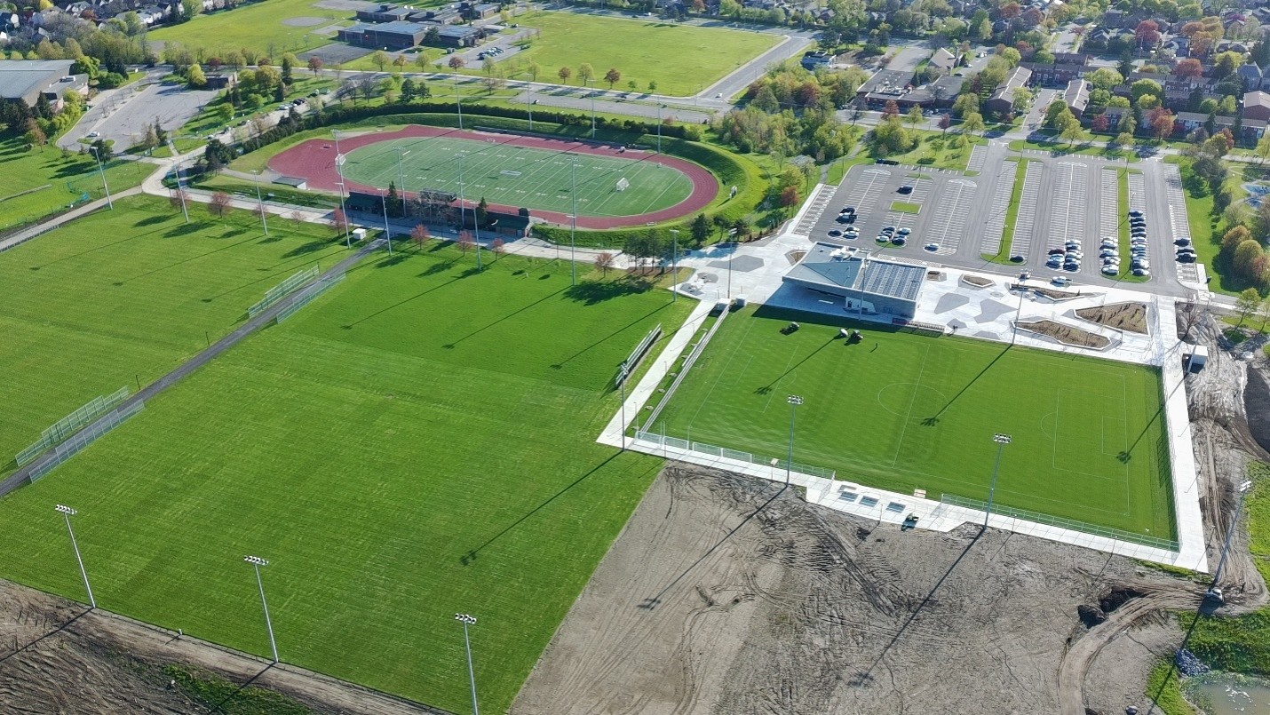 Aerial view of a sports complex featuring soccer fields and an athletics track around a sports field. New pathways connect the fields to a modern building and parking lot. Tall light poles are installed throughout the site.
