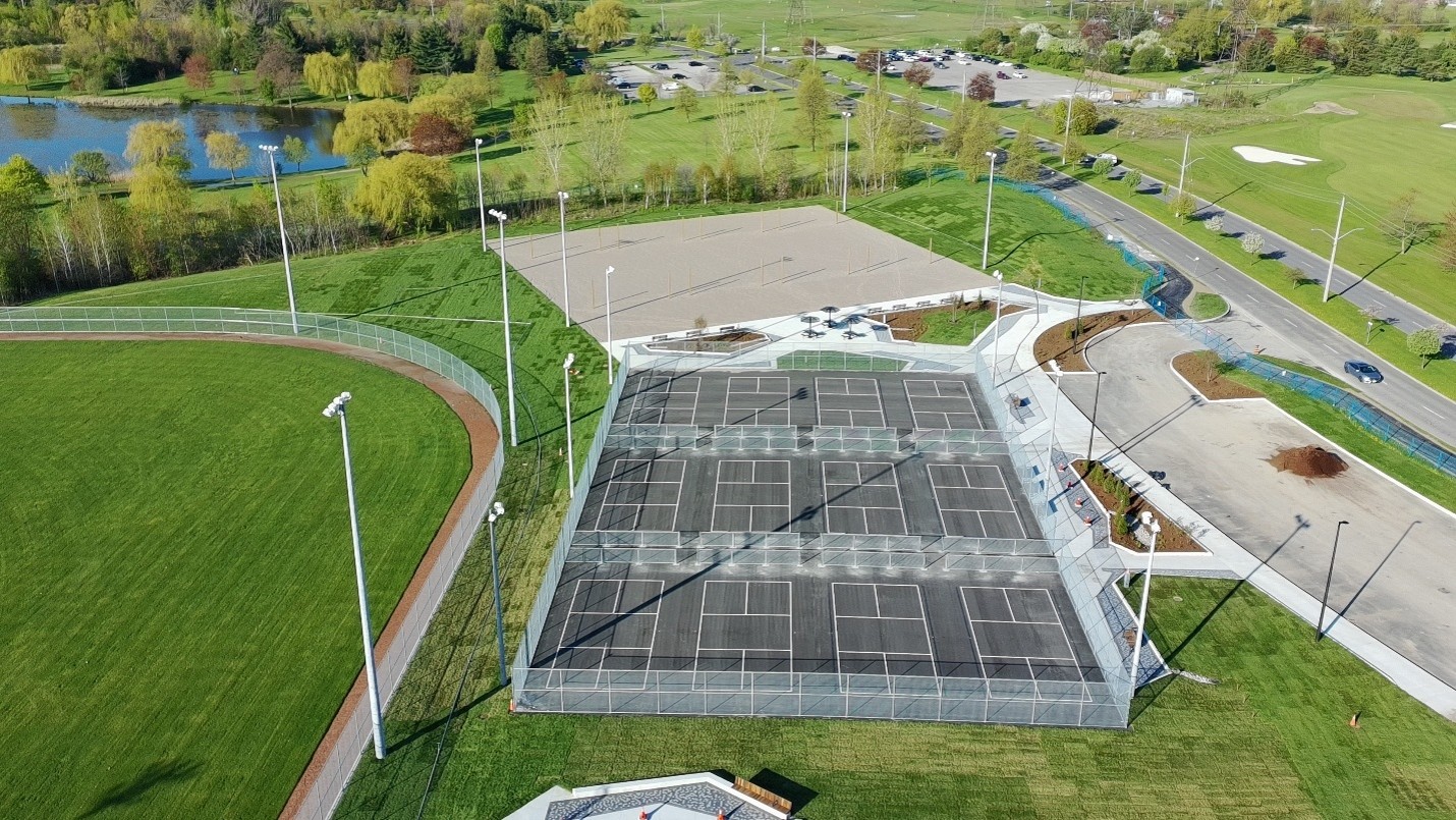 Aerial view of a sports complex featuring fenced pickleball courts beside volleyball courts. Multiple pickleball courts are arranged in rows, surrounded by pathways, seating areas, landscaping and tall light poles. A road, parking lots, ponds and mature trees are visible in the surrounding parkland.