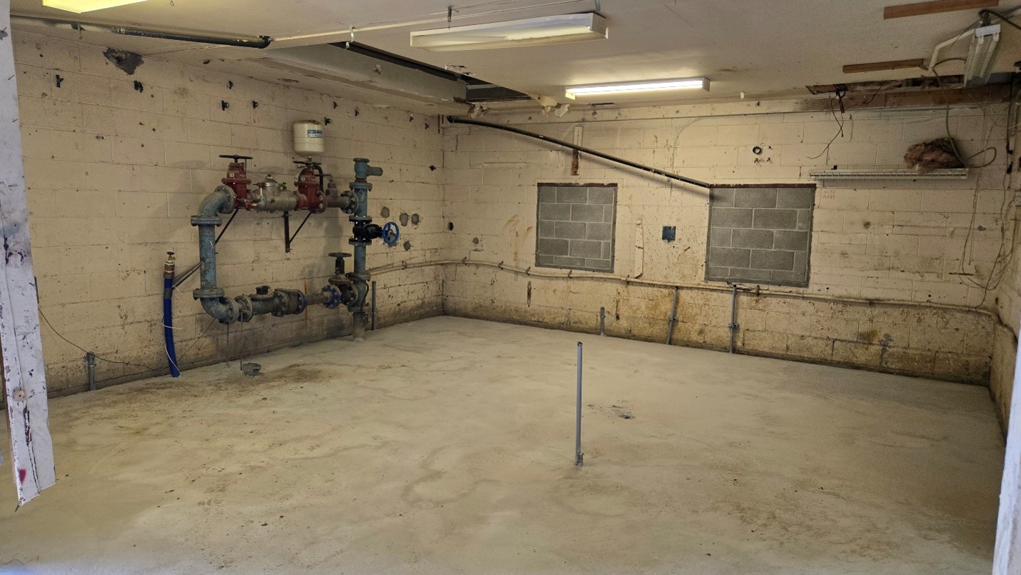Interior view of a room with block walls, concrete floor, exposed water pipes and electrical wiring. Fluorescent light fixtures are installed in the ceiling.