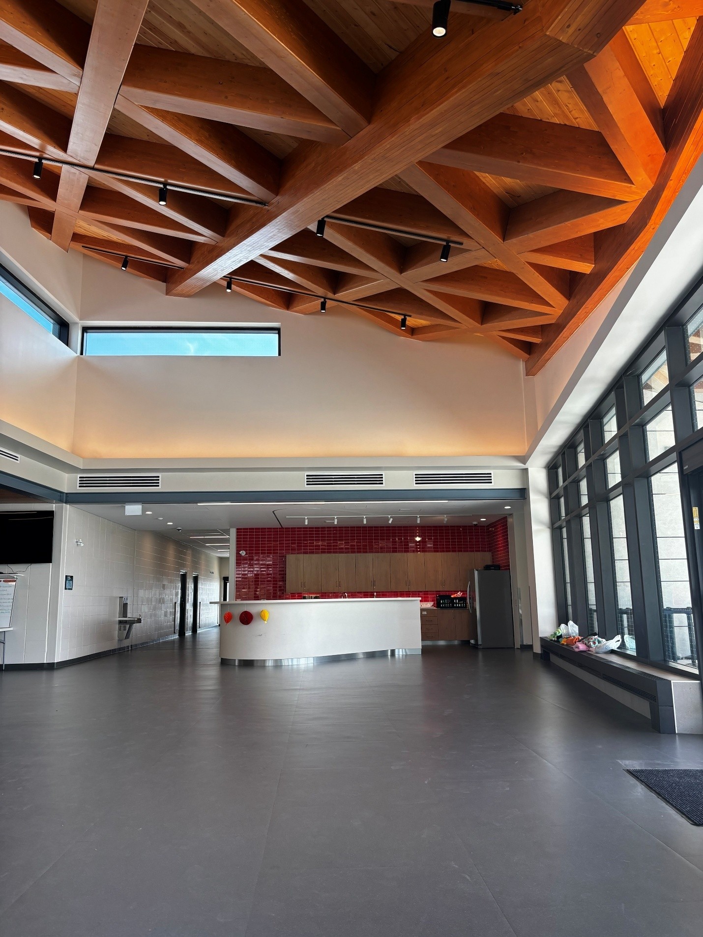 Interior view of a building featuring an open space with floor-to-ceiling windows, a reception counter and kitchenette area. The room has a high ceiling with exposed timber beams arranged in a geometric pattern.