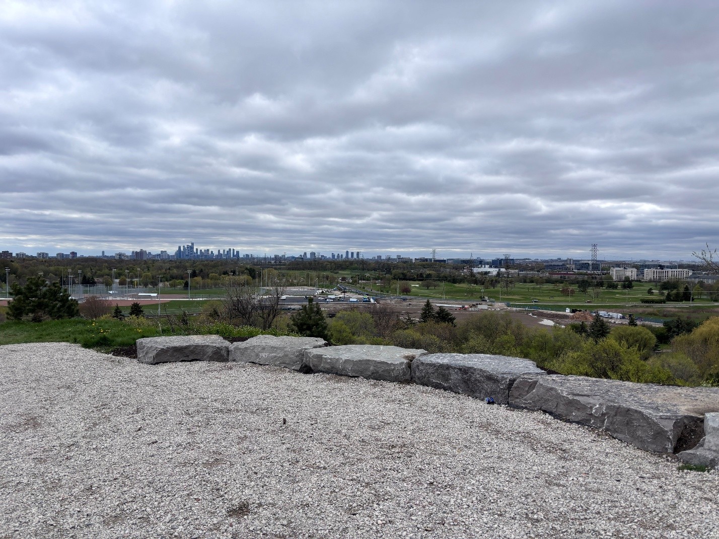 View from an elevated lookout over a large park. In the foreground, a gravel viewing area is bordered by large stone blocks. Beyond the lookout are open green spaces, sports fields and pathways. A city skyline is visible in the distance.